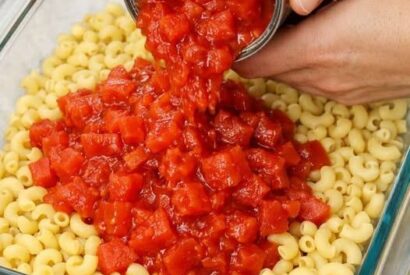Thumbnail for Layer raw elbow macaroni with canned diced tomatoes and two basic ingredients in a glass casserole dish for a cozy, crowd-pleasing dinner.
